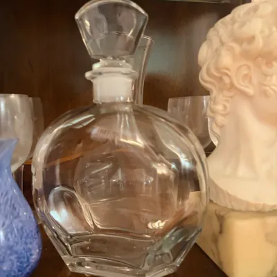 Clear glass decanter with a faceted glass stopper, displayed alongside glassware and a small classical bust on a wooden shelf.