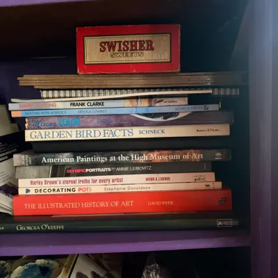 Stack of art and nature books, including works by David Piper and Annie Leibovitz, alongside a vintage Swisher Sweets cigar box on a...