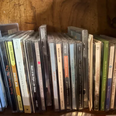 Sale Photo Thumbnail #207: A collection of music CDs in jewel cases stacked vertically on a wooden shelf, featuring various genres including jazz, blues, and relaxation music.