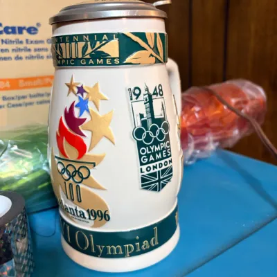 Sale Photo Thumbnail #220: Commemorative 1996 Atlanta Centennial Olympic Games beer stein with a metal lid and raised graphics representing historical Olympic host cities.