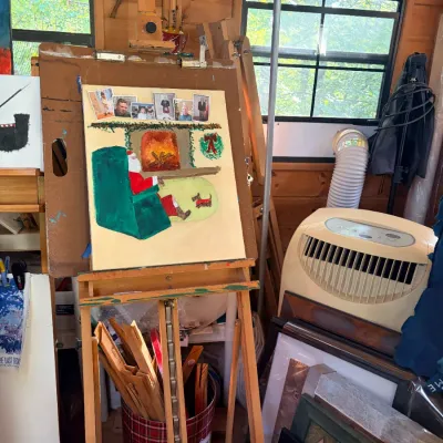 Wooden floor easel displaying an unfinished Christmas-themed painting, accompanied by various art supplies, picture frames, and a...