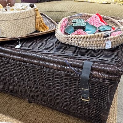 Large dark brown wicker trunk with leather strap closures, topped with decorative woven trays, a straw hat box, and teal ceramic dishes.
