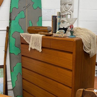 Sale Photo Thumbnail #26: Mid-century Danish modern teak tallboy dresser displaying tiki decor items, flanked by a vintage green and gray surfboard.