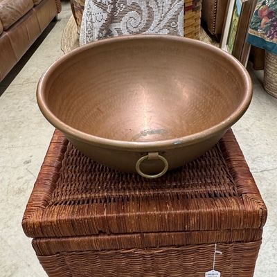 Large copper basin with ring side handles resting on a woven wicker storage trunk.
