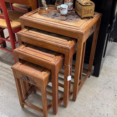 Sale Photo Thumbnail #18: Set of four hand-carved Asian wooden nesting tables featuring relief-carved top panels, displayed with decorative tusk accents and small porcelain pieces.
