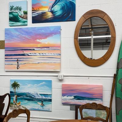 Sale Photo Thumbnail #25: Collection of original ocean and wave paintings hung on a wall alongside a oval wooden mirror, wooden dining set, and colorful fish-patterned ceramic platter.