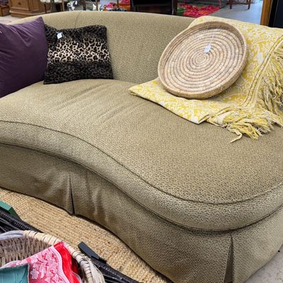 Sale Photo Thumbnail #22: Curved contemporary upholstered chaise lounge in textured olive green fabric, styled with accent pillows, a woven straw tray, and a yellow fringe throw blanket.