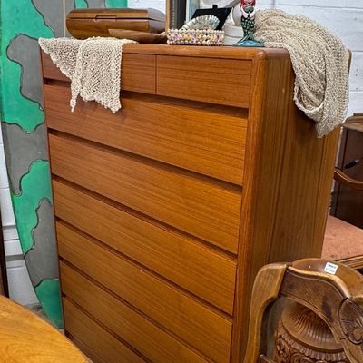 Danish Modern teak tallboy dresser featuring recessed drawer pulls, displayed with decorative decor alongside a surfboard and carved tiki...