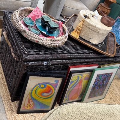 Sale Photo Thumbnail #20: Dark brown wicker trunk used as a coffee table, decorated with decorative trays, ceramic dishes, a woven hat box, and framed artwork leaning against the side.