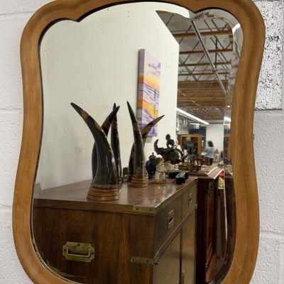Vintage wall mirror with a scalloped light wood frame mounted on a white painted cinderblock wall.