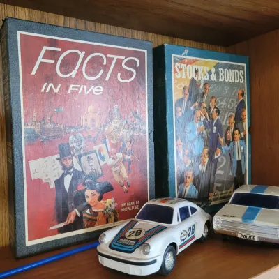 Sale Photo Thumbnail #29: Two 1960s 3M bookshelf games, Facts in Five and Stocks & Bonds, displayed on a wooden shelf alongside two vintage toy cars, including a Martini Porsche.
