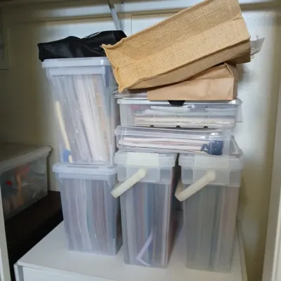Stack of plastic craft storage containers filled with paper and vinyl supplies, topped with a jute tote bag and paper bags.