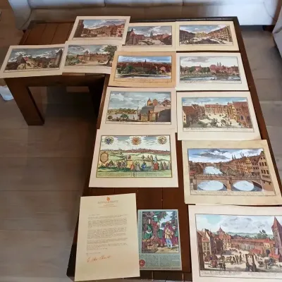 Collection of vintage hand-colored European city and landscape art prints laid out on a dark wood coffee table.