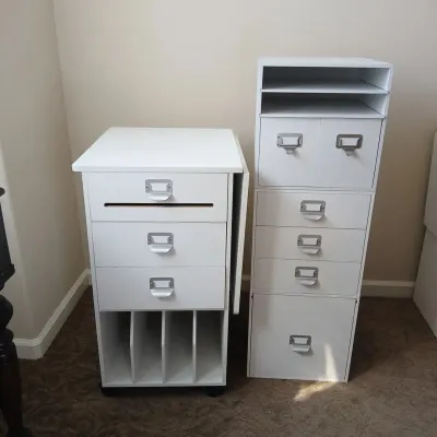Two white craft organization storage towers featuring multi-drawer units, open shelves, vertical paper slots, and silver label cup pulls.