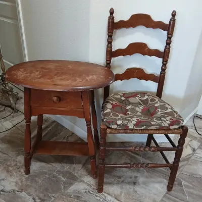Vintage wooden oval side table with a single drawer alongside a ladder-back chair featuring a rush seat and patterned cushion, showing...