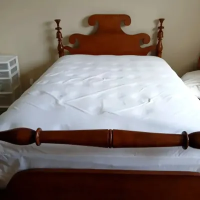 Vintage wooden bed frame featuring a scrolled crest headboard and turned finial posts, dressed in plain white bedding.