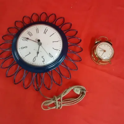 Vintage blue metal Westclox electric sunburst wall clock alongside a small brass twin-bell alarm clock, resting on a red surface.