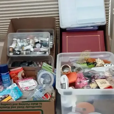 Large assortment of crafting supplies, including ribbons, tape, glitter jars, and organizational plastic bins, stored in U-Haul boxes.