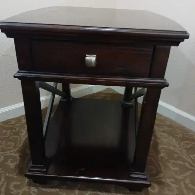 Dark wood end table featuring a single drawer with a brushed metal knob, lower shelf, and crossed side supports.