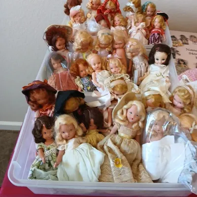 Large collection of vintage storybook and character dolls, including Madame Alexander and similar styles, displayed in plastic storage bins.