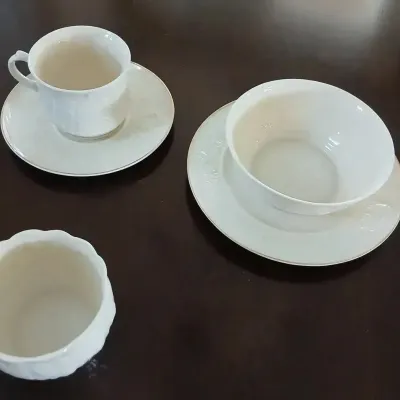 White porcelain tableware set featuring subtle embossed floral trim and gold-tone rims, including a teacup with saucer, a small bowl on a...