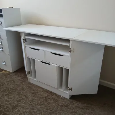 White craft and sewing cabinet featuring a fold-out tabletop extension, multiple drawers, and open storage slots.