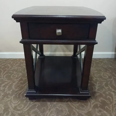 Dark wood end table featuring a single drawer with a brushed metal pull, lower display shelf, and decorative X-brace side supports.