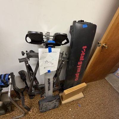 Murtisol folding rowing machine with user manual alongside a Total Gym Pro bench, folded and stored against a wall.