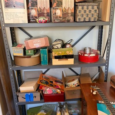 Metal shelving unit filled with vintage boxed lamps, craft kits, sewing supplies, and tin containers, alongside a wooden desk with a banjo.