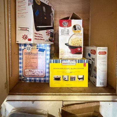 Cabinet shelf filled with boxed kitchen appliances, including a George Foreman grill, a West Bend popcorn machine, a Toastmaster bread...