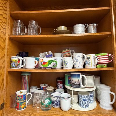 Wooden kitchen cabinet filled with an eclectic collection of vintage and souvenir coffee mugs, glassware, and aluminum measuring cups.