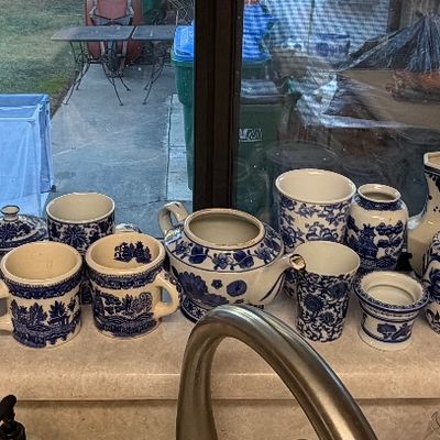 Collection of blue and white ceramic items, including various pitchers, vases, mugs, and a teapot, displayed along a window sill.