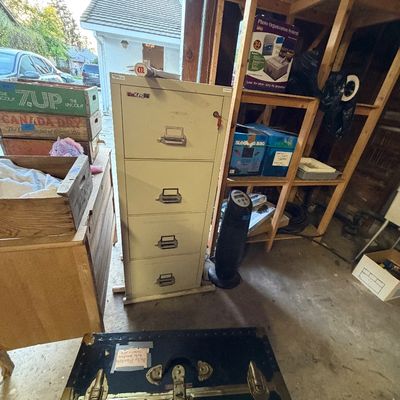 Vintage FireKing four-drawer vertical file cabinet in beige metal, set in a garage with wooden crates, a space heater, and a blue trunk...