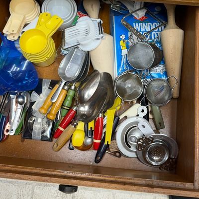 Wooden kitchen drawer filled with various vintage utensils, including metal scoops, strainers, measuring cups, and an egg slicer.