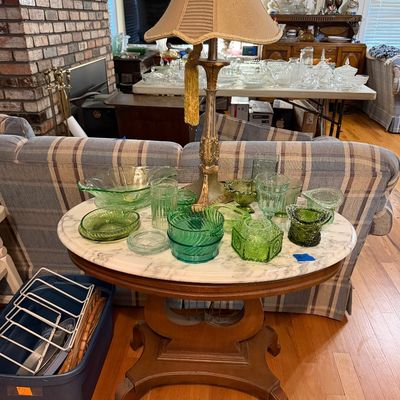 Collection of vintage green depression glass and glassware displayed on a round marble-top pedestal side table with a brass lamp.