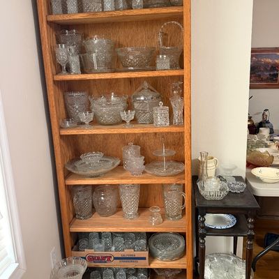 Tall wooden bookcase and small side table displaying a large collection of clear pressed and cut glassware, including bowls, pitchers,...