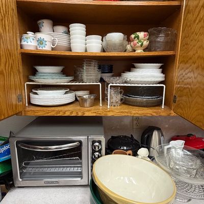 Kitchen cabinet filled with assorted glassware, ceramic ramekins, floral teacups, and dinnerware, alongside a stainless steel KitchenAid...