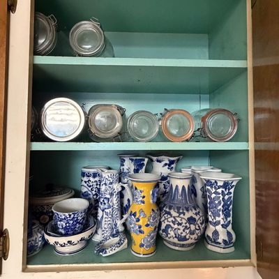 A turquoise painted cabinet filled with a collection of blue and white ceramic vases, tea sets, and a yellow floral vase, alongside glass...