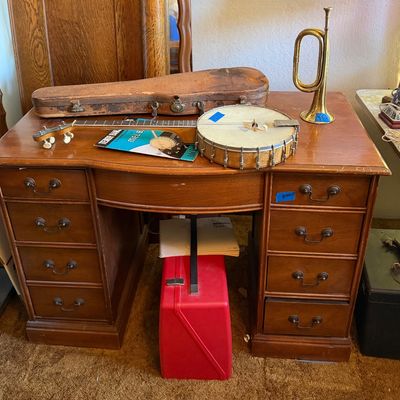Vintage wooden kneehole desk featuring musical instruments on top, including a banjo with case, sheet music, and a brass bugle.