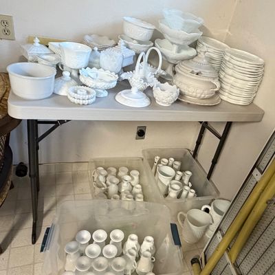 Large collection of white milk glass tableware, including covered dishes, plates, footed bowls, and drinkware, displayed on a folding...