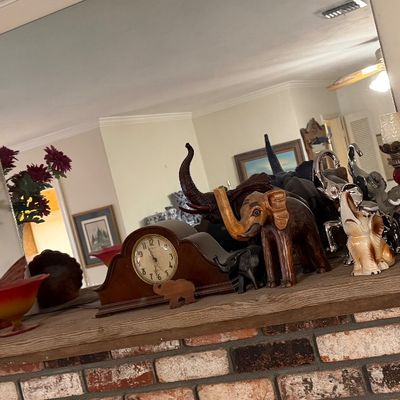 Collection of decorative elephant figurines, a wooden mantle clock, and a pink Fenton hobnail glass parlor lamp arranged on a brick...