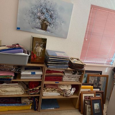 Sale Photo Thumbnail #107: Collection of folded linens, fabric remnants, and photo albums displayed on light wood bookcases, alongside framed wall art and decor.