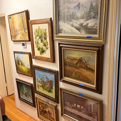 Gallery wall featuring a variety of framed vintage art pieces, including landscape oil paintings, embroidered textile art, and a print of...