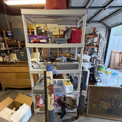 White plastic four-tier shelving unit holding red gas cans, toolboxes, and miscellaneous garage items, shown alongside framed still life...