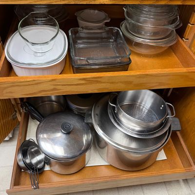 Wooden cabinet filled with vintage glass bakeware, Pyrex cookware, and stainless steel pots and pans.