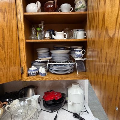 Kitchen cabinet filled with ceramic tableware and teapots, including blue and white transferware, alongside small appliances on the...