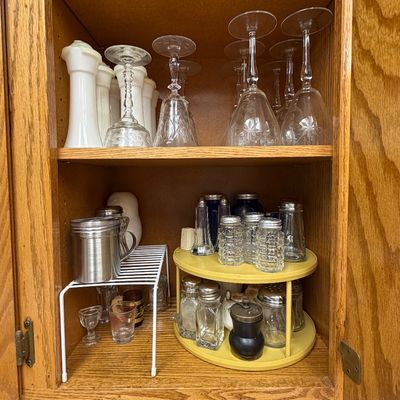 Wooden kitchen cabinet filled with vintage etched glassware, milk glass vases, and a yellow Lazy Susan organizing salt and pepper shakers.