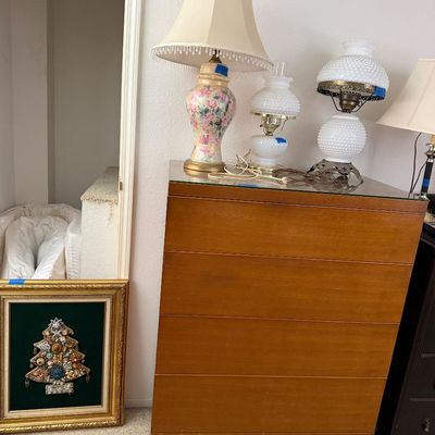 Sale Photo Thumbnail #109: Mid-century wooden tallboy dresser with a glass top, featuring several vintage table lamps and hobnail glass oil-style lamps on top, alongside framed jewelry tree artwork on the floor.