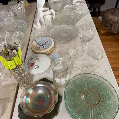 Collection of vintage glassware and porcelain tableware displayed on a folding table, including press glass platters, carnival glass...