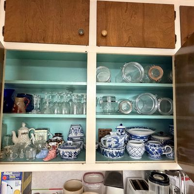 Open wooden kitchen cabinet painted turquoise inside, filled with various glassware, storage jars, and blue-and-white porcelain tea sets.